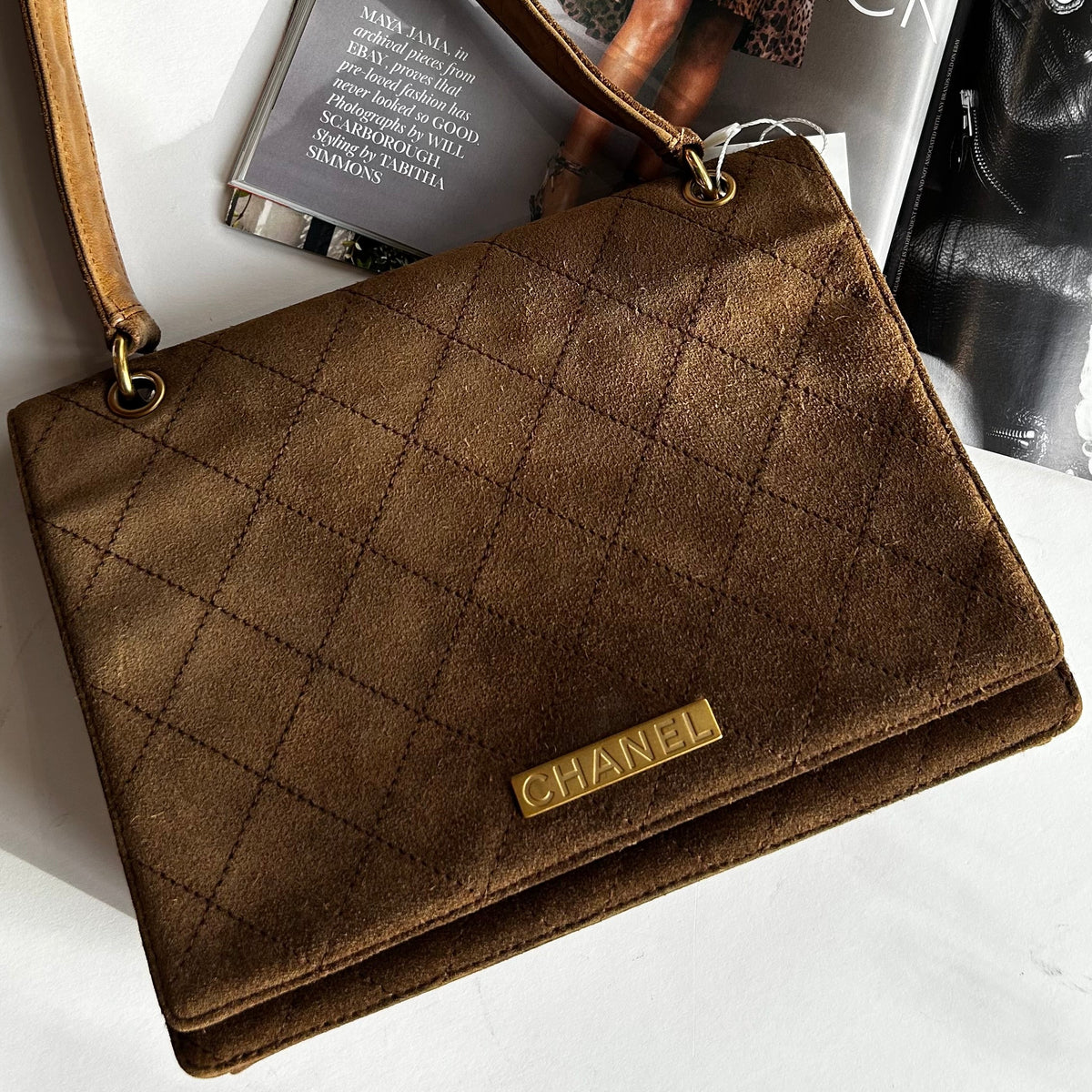 Pre-loved Brown quilted Chanel handbag on a white surface with a magazine in the background. 