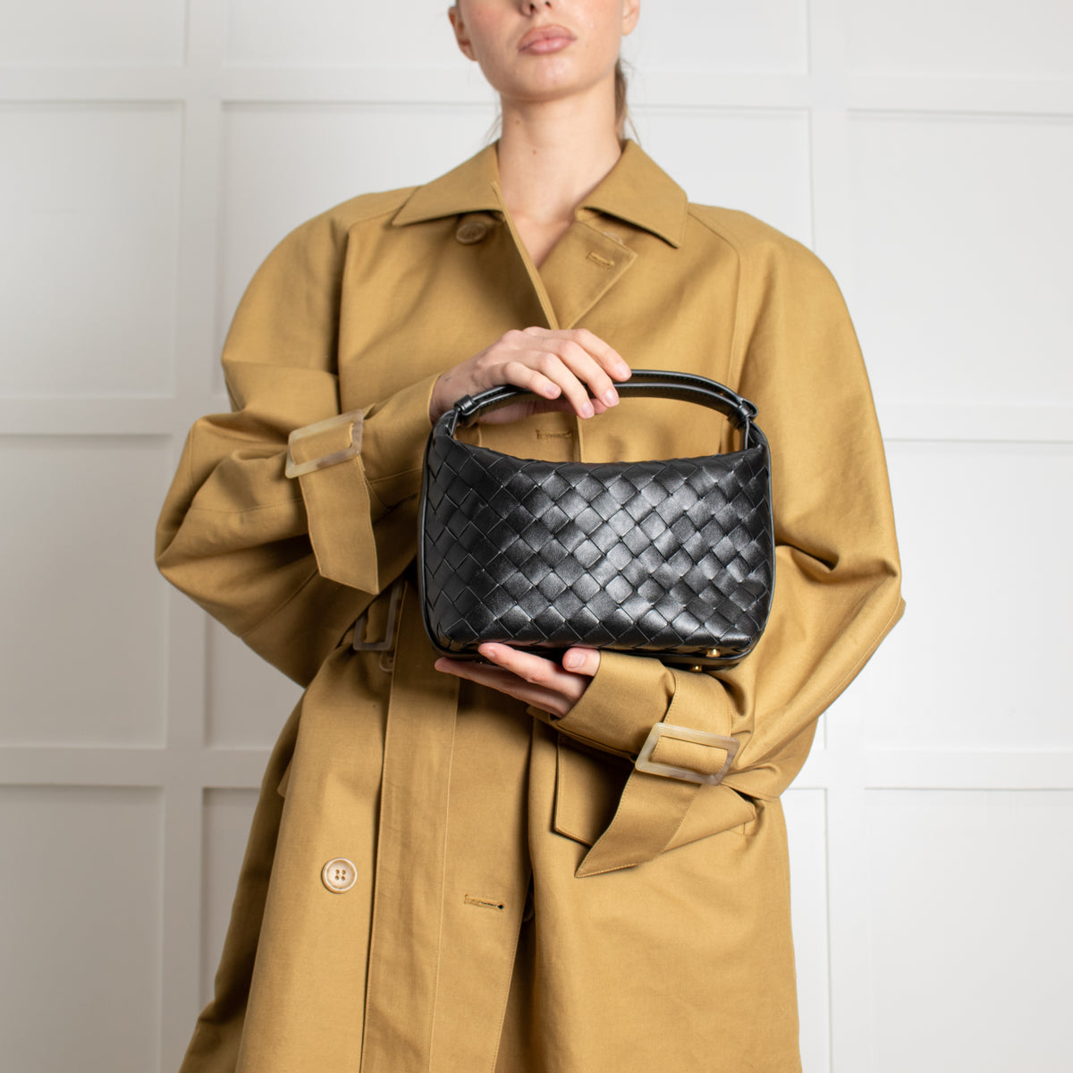 Person wearing a tan trench coat holding a black preloved Bottega woven handbag against a white background.