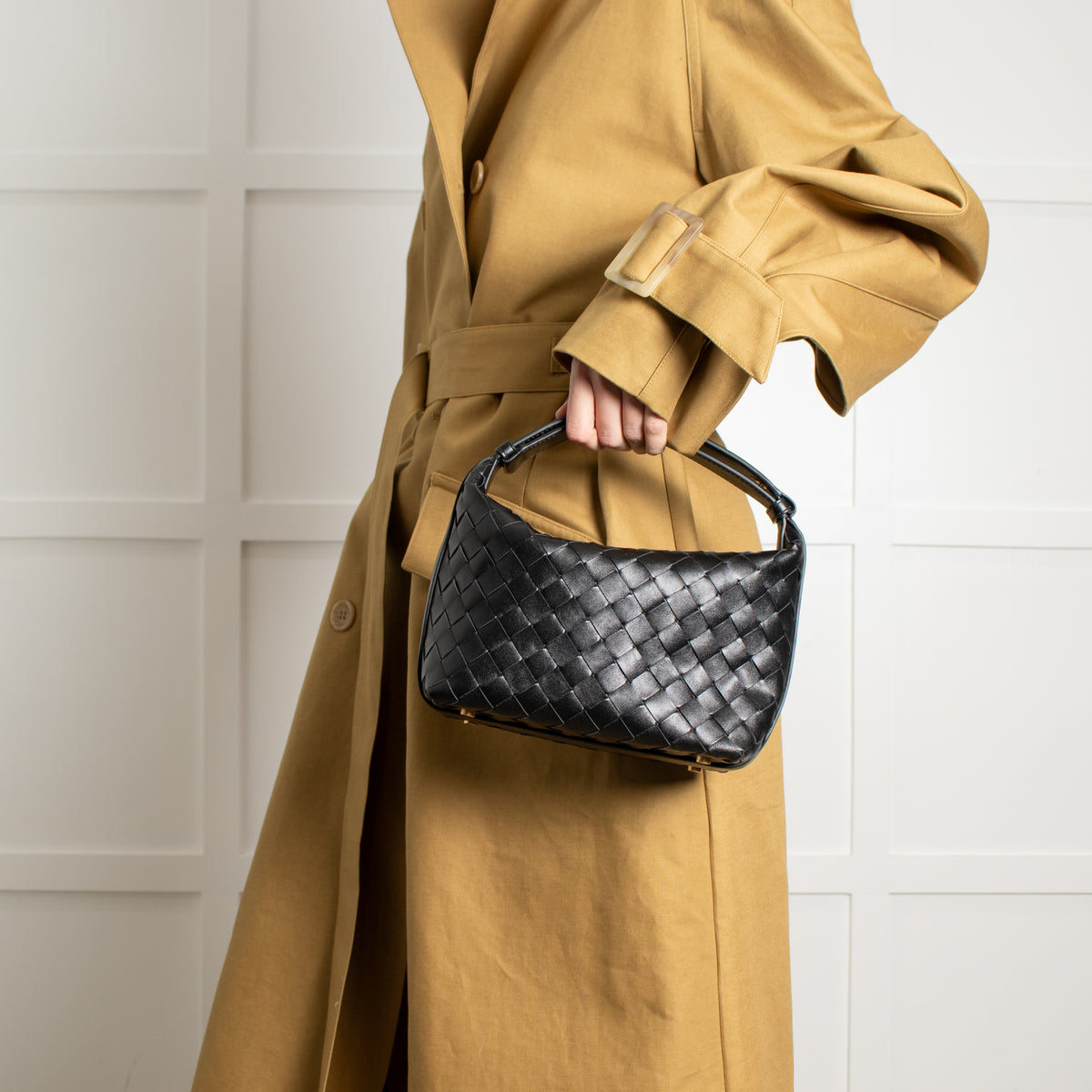 Person wearing a tan trench coat holding a black Bottega Veneta woven handbag against a white wall.