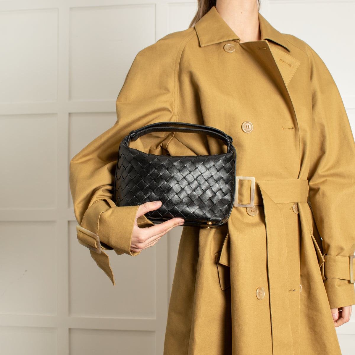 Model wearing a tan trench coat holding a black woven Bottega Veneta handbag against a neutral background. 