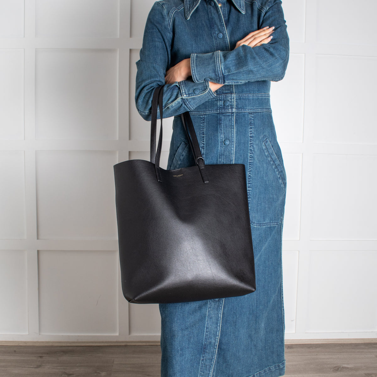 Person wearing a denim outfit holding a black Saint Laurent tote bag against a white paneled wall.