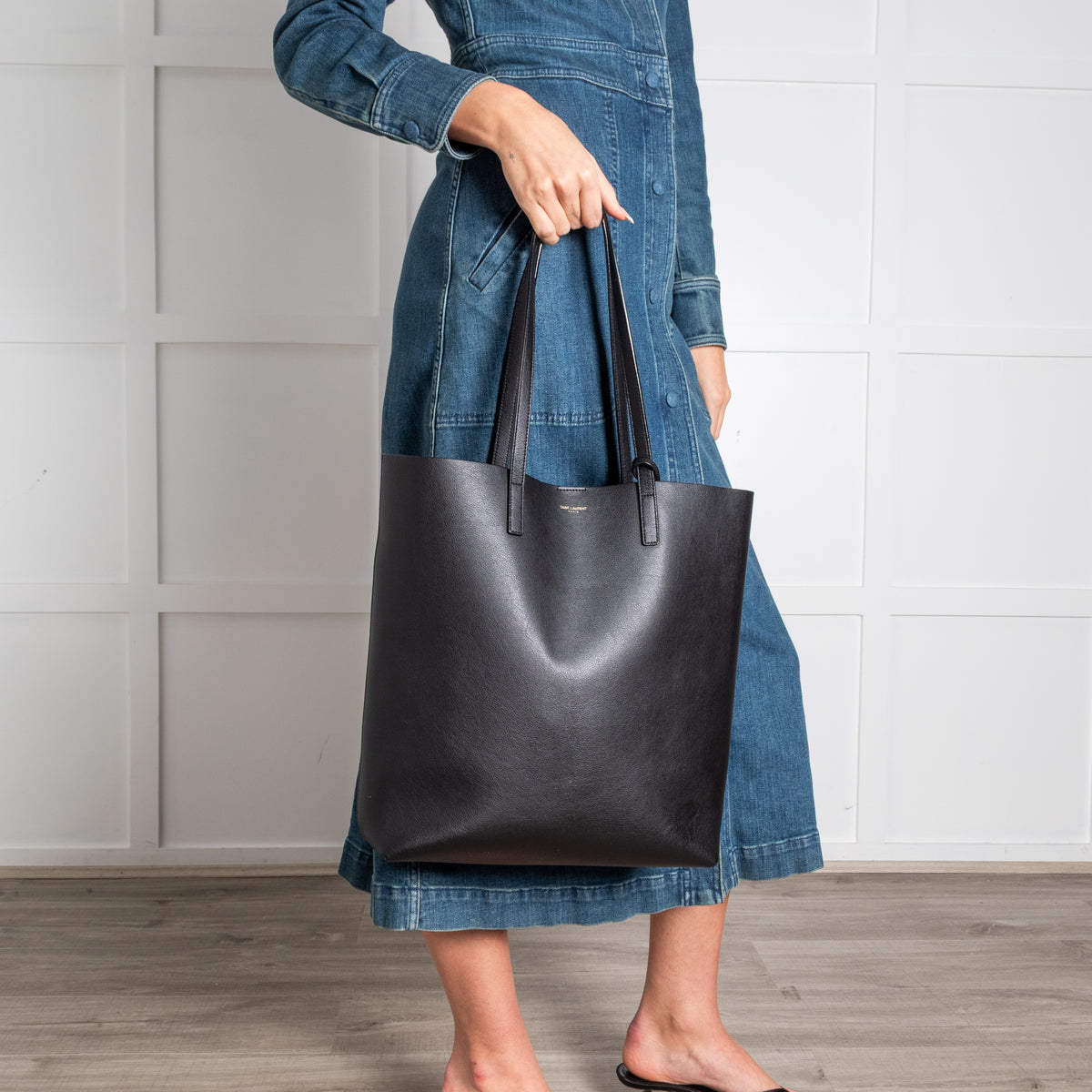 Person holding a black Saint Laurent leather tote bag against a white wall.